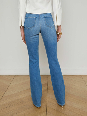 A person stands on a wooden floor, shown from the waist down wearing L'AGENCE Sale's Selma Bootcut Jean in blue and a white long-sleeve top featuring gold zipper accents on the sleeves.