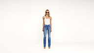 A woman wearing sunglasses, a white tank top, blue jeans, and heels stands against a plain white background, facing forward with arms relaxed at her sides.