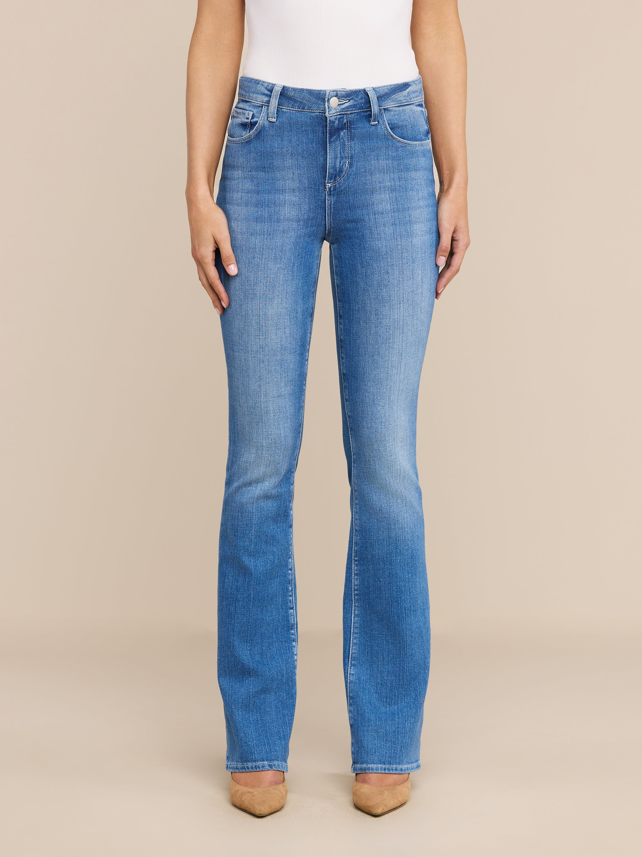 Una donna rivolta in avanti indossa il Selma Bootcut Jean in denim elasticizzato a lavaggio medio, un top bianco senza maniche e scarpe beige a punta, su uno sfondo beige neutro.