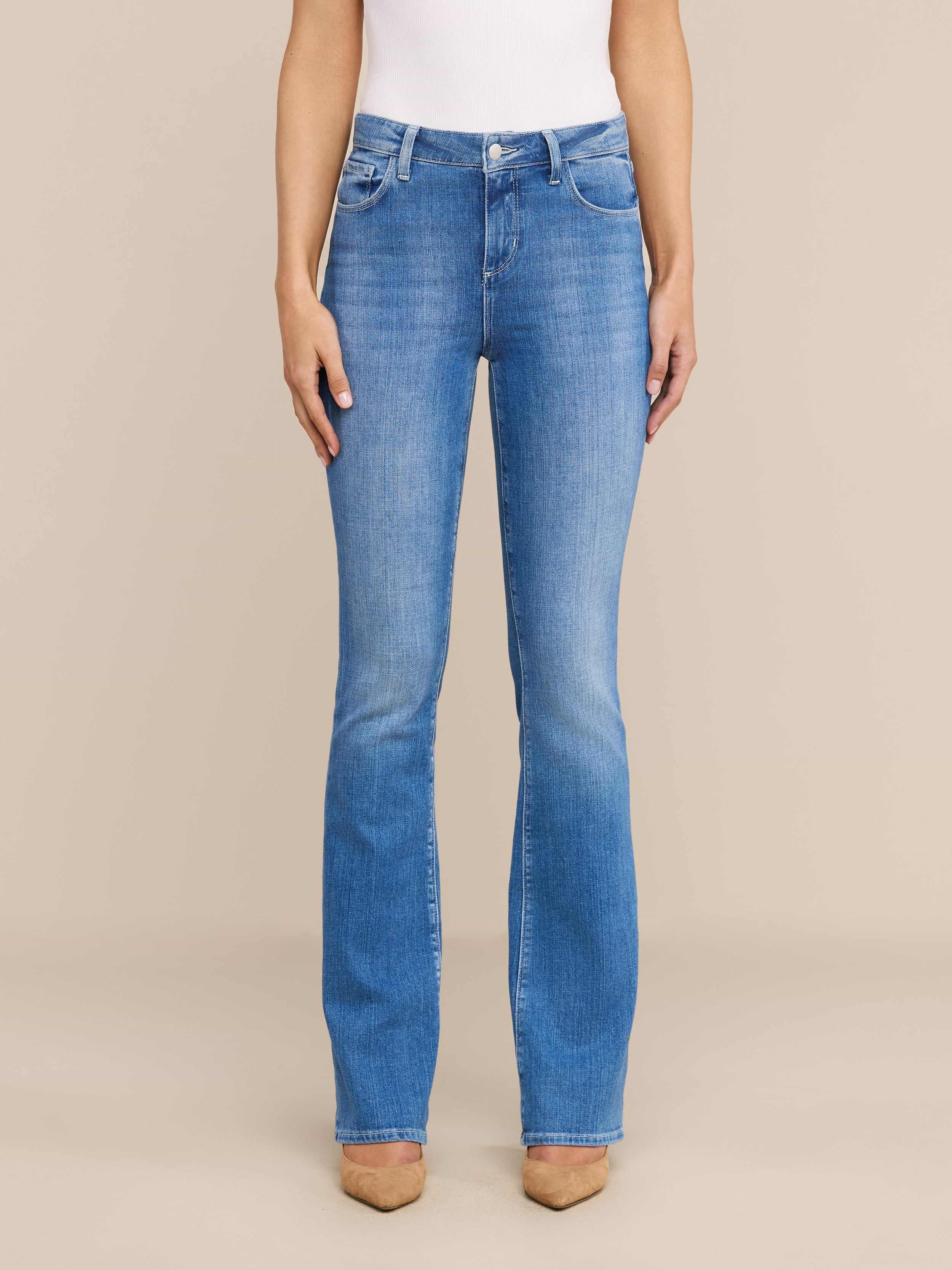Una donna rivolta in avanti indossa il Selma Bootcut Jean in denim elasticizzato a lavaggio medio, un top bianco senza maniche e scarpe beige a punta, su uno sfondo beige neutro.