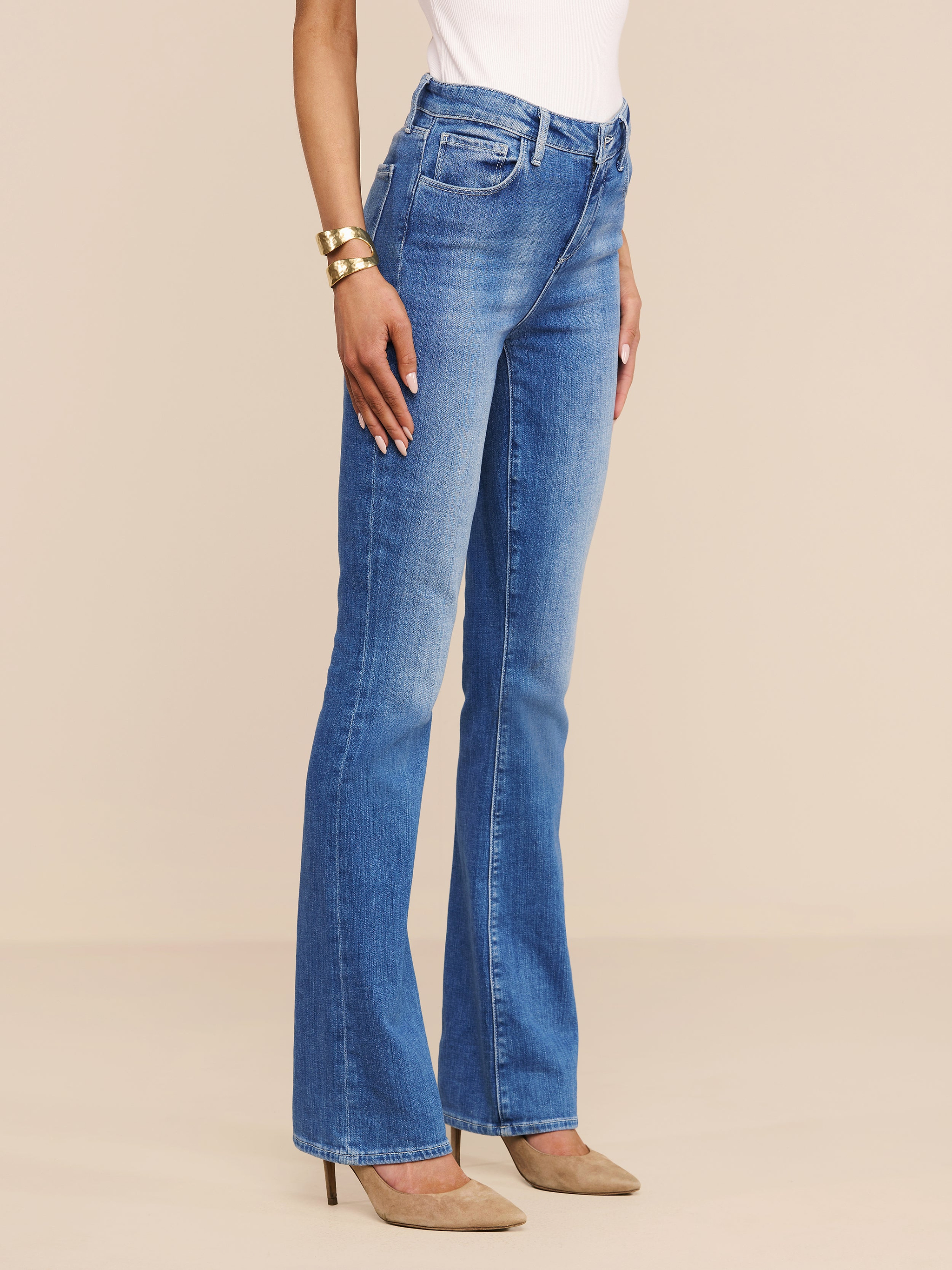 Una persona indossa Selma Bootcut Jean in blu con un top bianco, tacchi alti marrone e braccialetti doro, su uno sfondo beige.