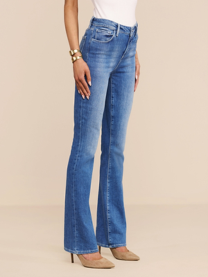 A person wears Selma Bootcut Jean in blue with a white top, tan high heels, and gold bracelets, standing against a plain beige background.