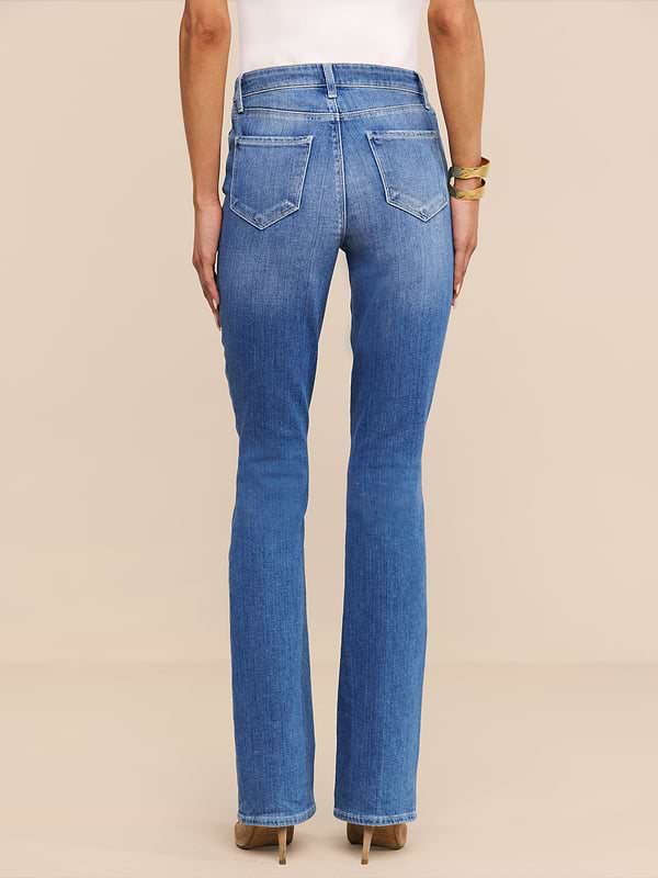 Vista di spalle su uno sfondo beige, una persona indossa il Selma Bootcut Jean in denim elasticizzato a lavaggio medio, abbinato a un top bianco, scarpe con tacco marrone e bracciali dorati.