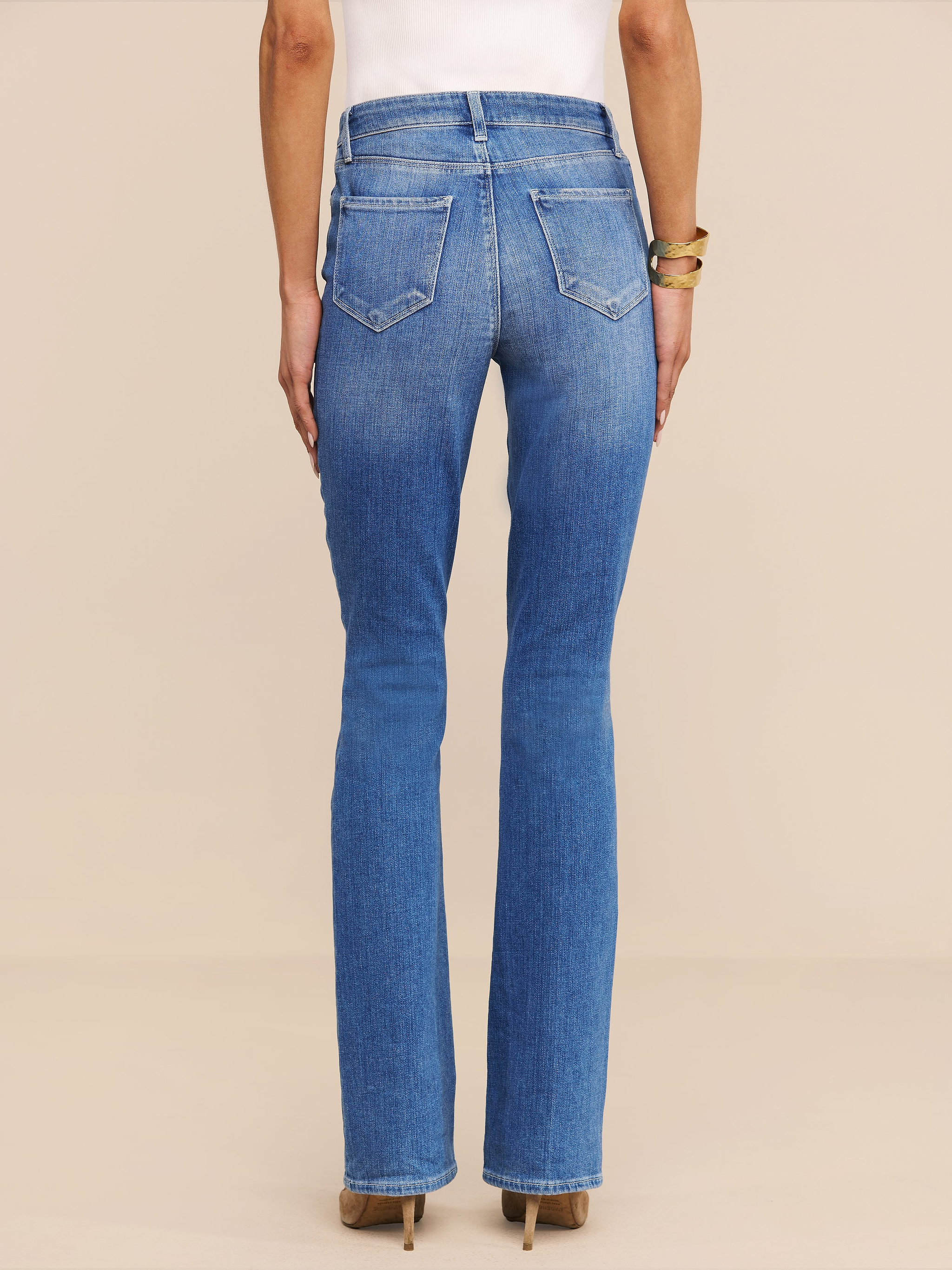 Vista di spalle su uno sfondo beige, una persona indossa il Selma Bootcut Jean in denim elasticizzato a lavaggio medio, abbinato a un top bianco, scarpe con tacco marrone e bracciali dorati.