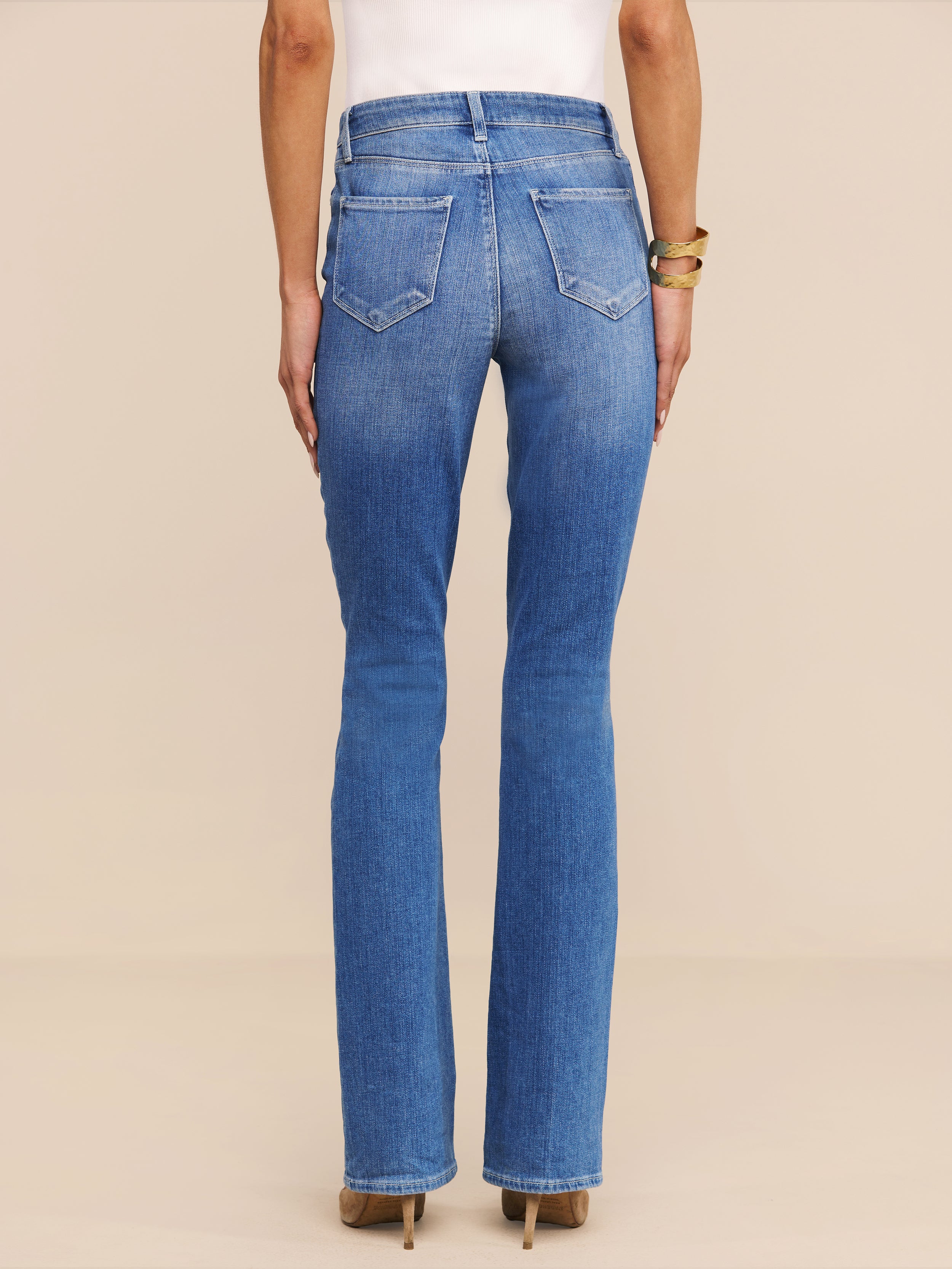 Vista di spalle su uno sfondo beige, una persona indossa il Selma Bootcut Jean in denim elasticizzato a lavaggio medio, abbinato a un top bianco, scarpe con tacco marrone e bracciali dorati.