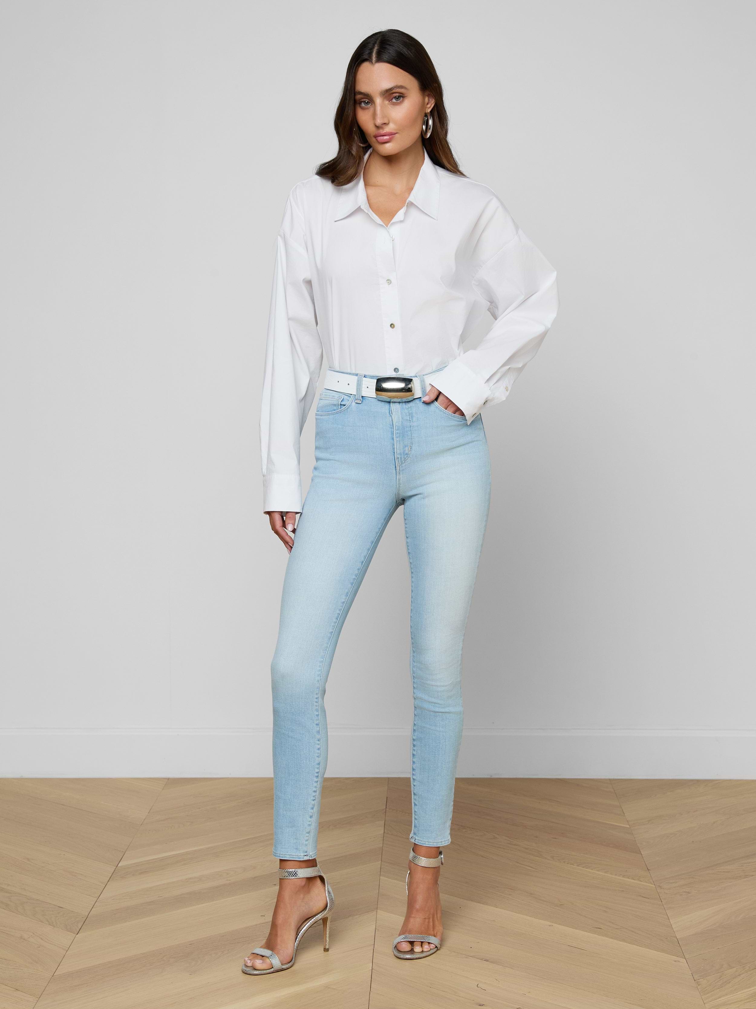 A woman poses indoors with one hand on her hip, wearing a loose white button-up shirt, the Monique Skinny Jean in light blue, a white belt, and silver high-heeled sandals against a plain background.