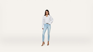 A woman stands against a plain white background, wearing a white button-up shirt, light blue skinny jeans, and beige high-heeled sandals. She has one hand on her hip and looks confidently at the camera.