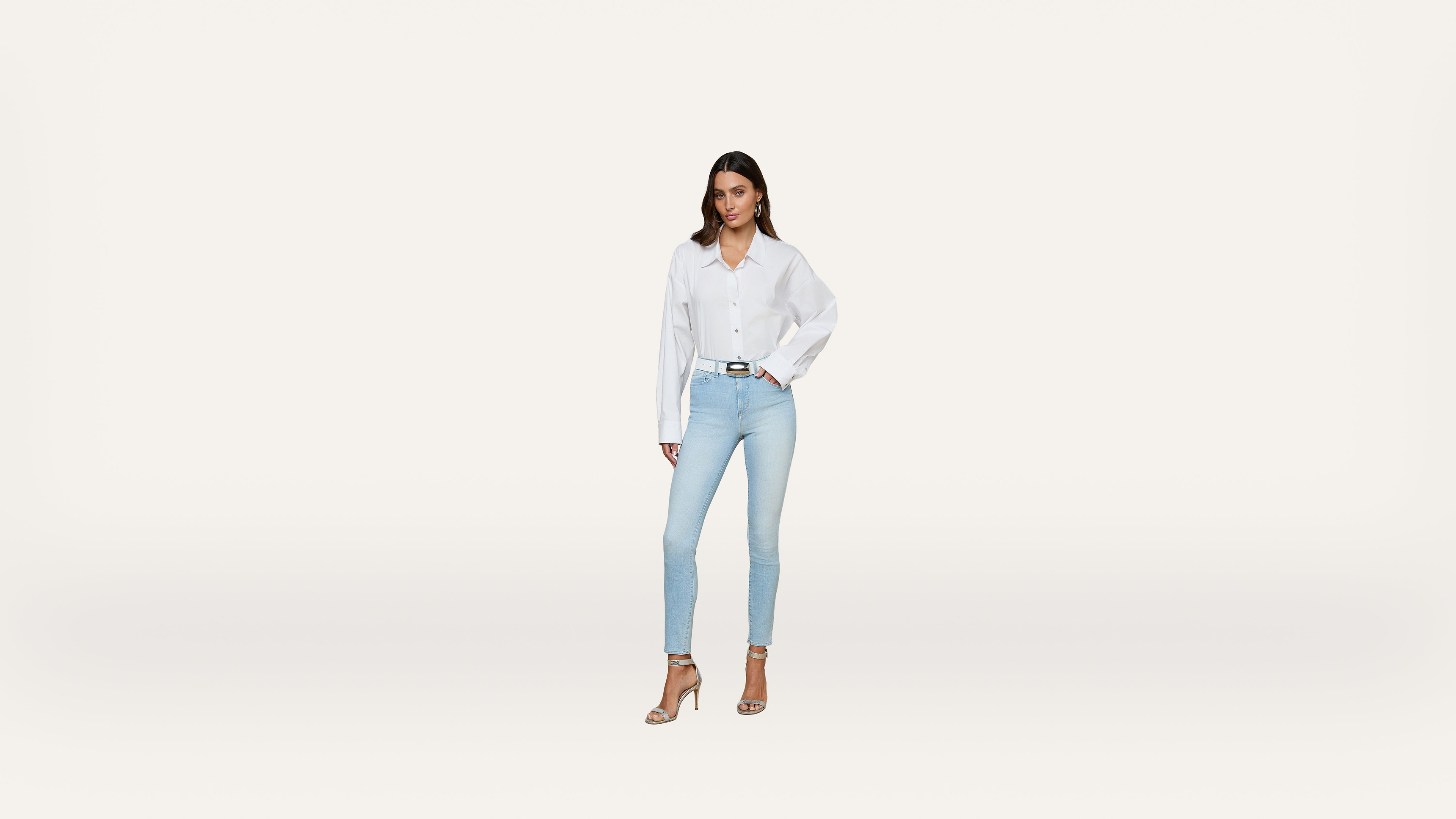 A woman stands against a plain white background, wearing a white button-up shirt, light blue skinny jeans, and beige high-heeled sandals. She has one hand on her hip and looks confidently at the camera.