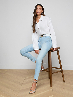 A woman with long brown hair sits on a wooden stool against a plain white background, wearing a white button-up shirt, a white belt, silver high-heeled sandals, and the Monique Skinny Jean crafted from stretch denim.
