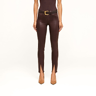 Wearing the Jyothi Coated Jean—high-waisted, brown, and featuring front ankle slits—a person is styled with a black belt with a gold buckle, a sleeveless dark top, and tan pointed-toe heels. The image shows them from the shoulders down against a white background.