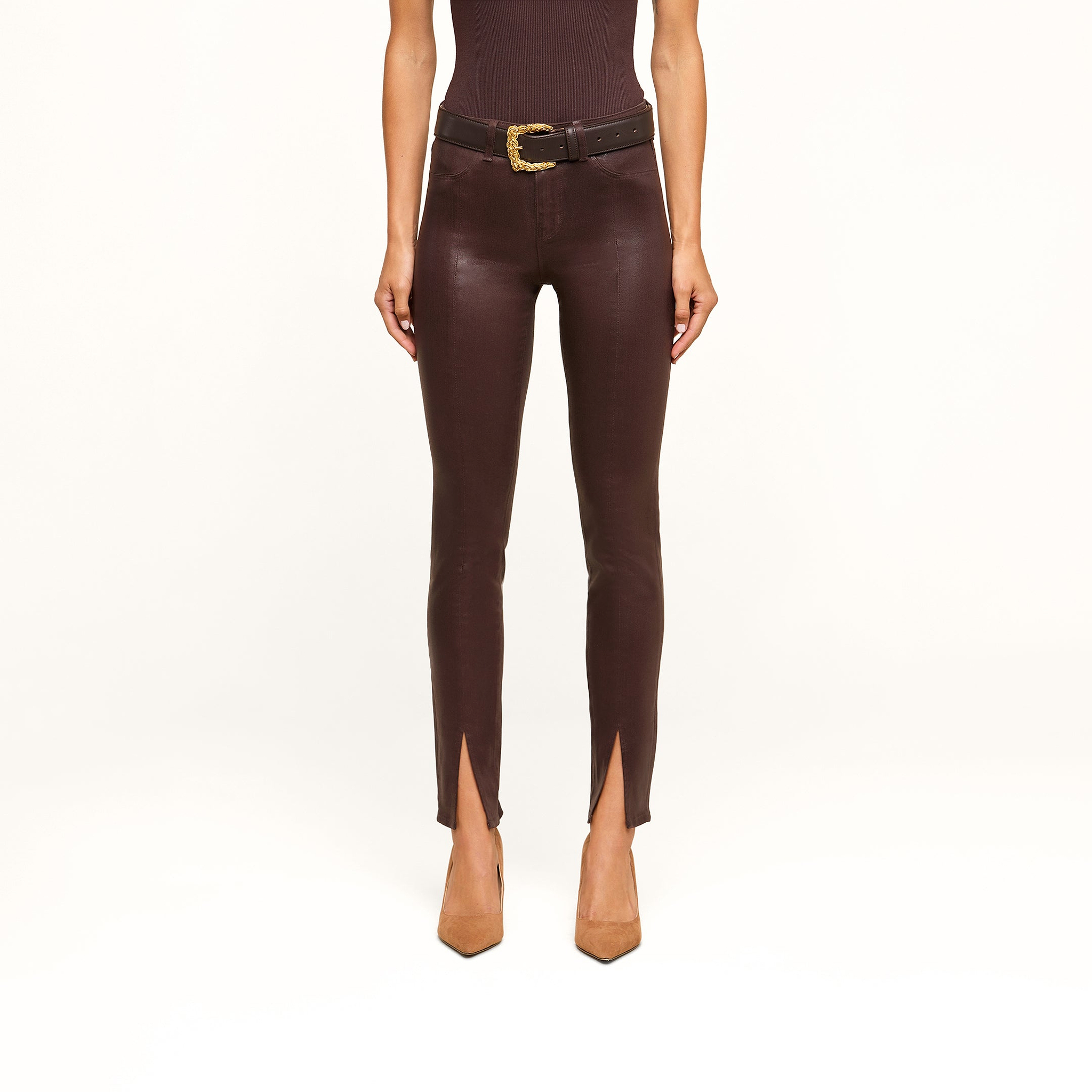 Wearing the Jyothi Coated Jean—high-waisted, brown, and featuring front ankle slits—a person is styled with a black belt with a gold buckle, a sleeveless dark top, and tan pointed-toe heels. The image shows them from the shoulders down against a white background.
