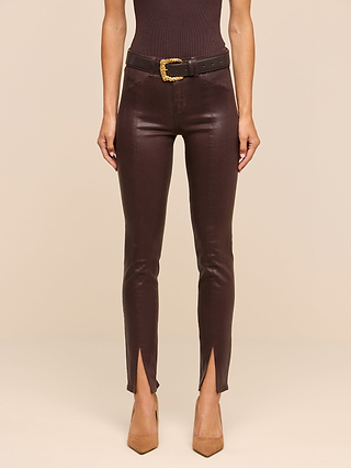 A woman stands against a beige background wearing the Jyothi Coated Jean—high-waisted, brown coated denim pants featuring front ankle slits—styled with a black belt with a gold buckle and tan pointed heels.