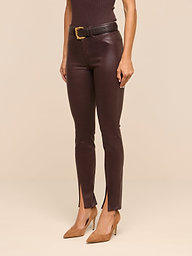 A woman wearing the Jyothi Coated Jean—brown high-rise skinny jeans with front ankle slits—paired with a black top, a black belt featuring a gold buckle, and tan pointed-toe heels, standing against a plain white background.