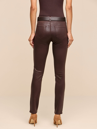 A person stands facing away, wearing a sleeveless dark brown top and the Jyothi Coated Jean—high-rise skinny jeans in fitted brown coated denim with a belt—paired with tan heeled shoes, against a beige background.