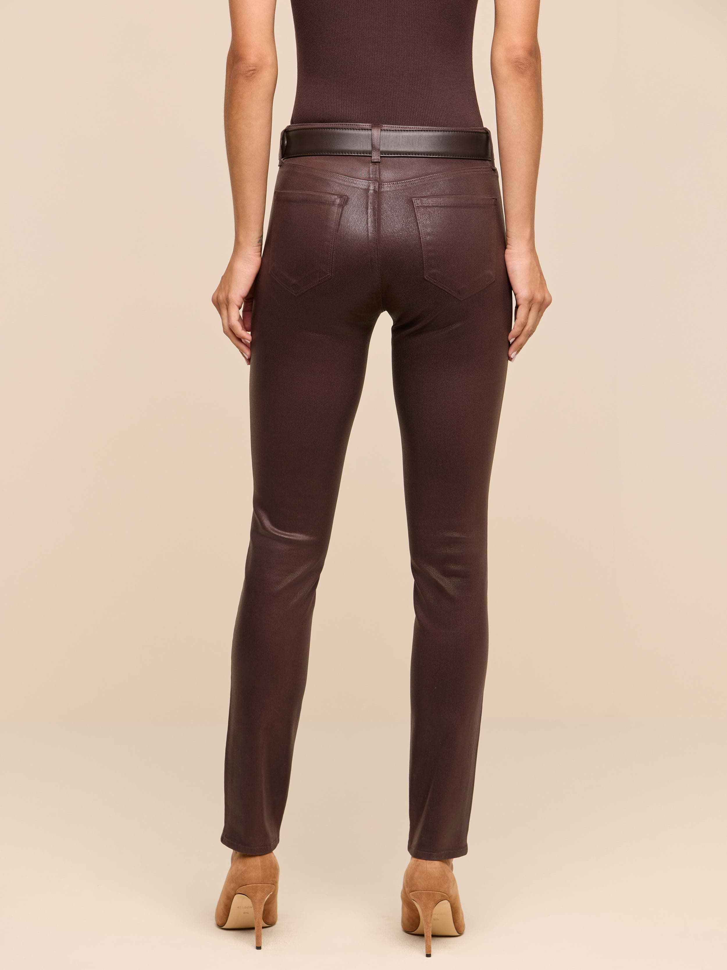 A person stands facing away against a plain white background, wearing a sleeveless brown top, the Jyothi Coated Jean in dark brown—featuring high-rise skinny fit with pockets and an ankle slit—a black belt, and tan high-heeled shoes.