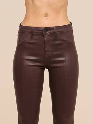 A close-up of a person wearing the Jyothi Coated Jean in shiny, dark brown, featuring a high-waisted fit with front pockets, button, and zipper. The background is beige.