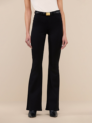 Someone stands against a beige background wearing the Marty Flare Pant—high-waisted, black ponte flare-leg pants with a black belt and gold rectangular buckle—paired with black shoes and a sleeveless white top.