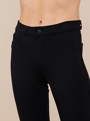 A close-up of a person wearing the Marty Flare Pant, a high-waisted black ponte slimming style with button and zipper closure, showing the waist and upper thigh area against a plain beige background.