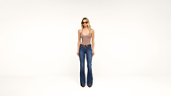 A woman wearing sunglasses, a tan halter top, blue flared jeans, and brown boots stands against a plain white background.