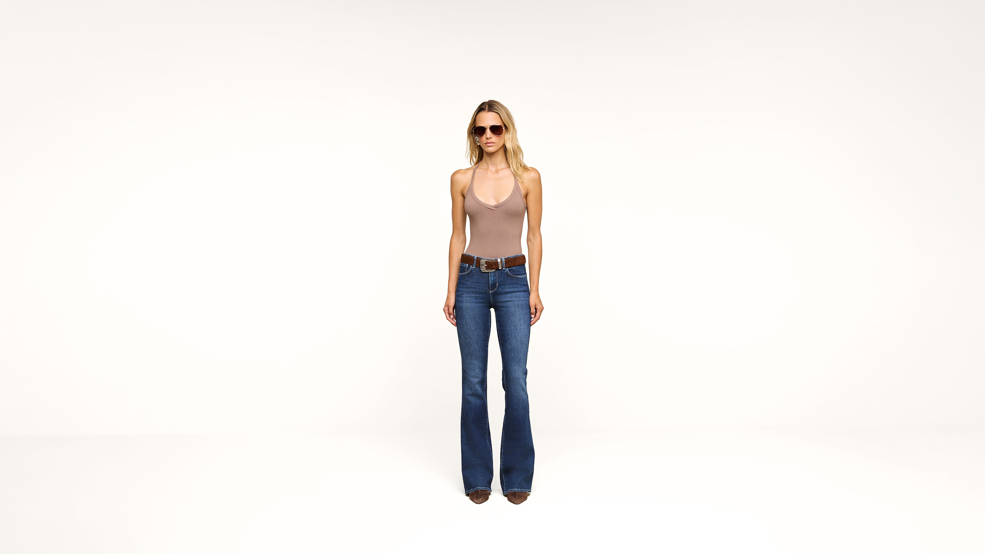 A woman wearing sunglasses, a tan halter top, blue flared jeans, and brown boots stands against a plain white background.