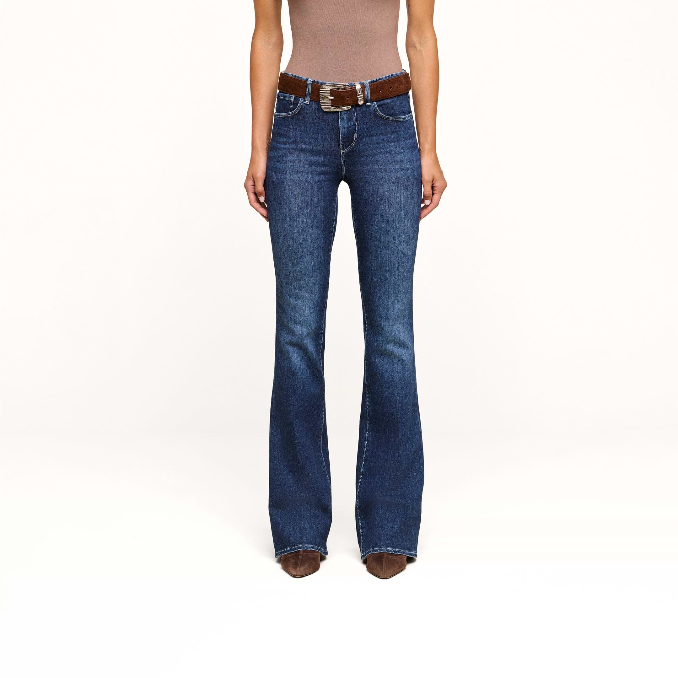 A person wearing a brown sleeveless top, the high-rise Marty Flare Jean crafted from stretch denim with a brown belt, and brown pointed-toe shoes stands against a plain white background.