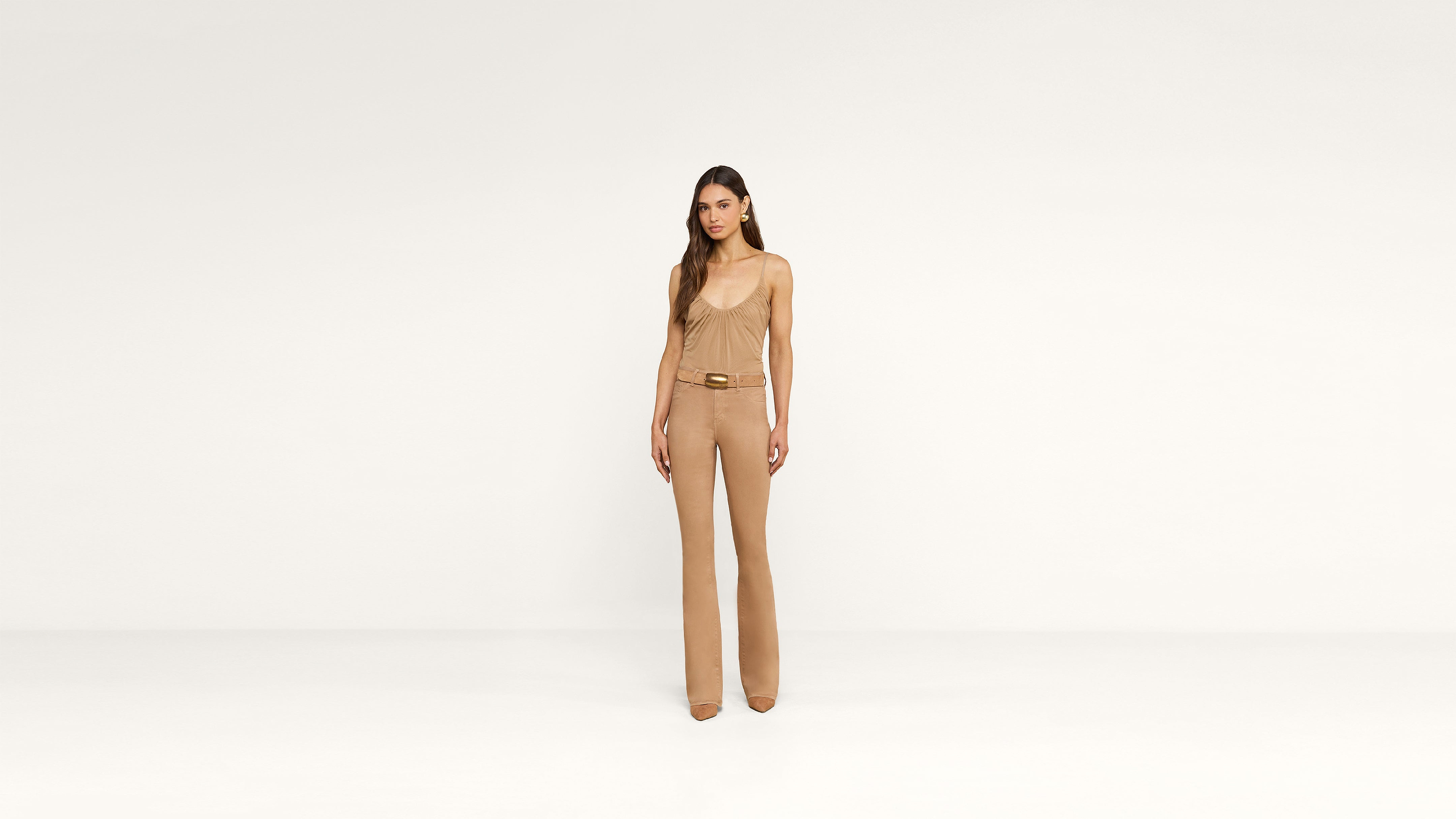 A woman stands in the center of a plain white background, wearing a tan sleeveless top, matching high-waisted pants with a belt, and tan heels. Her long hair falls loosely around her shoulders, complementing her neutral expression.