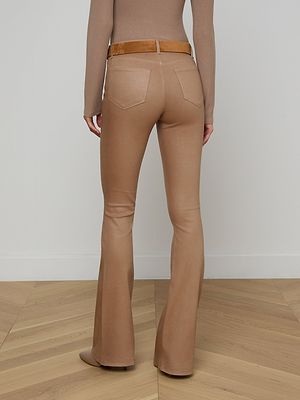 A person stands indoors on light wooden flooring against a white wall, wearing a fitted beige long-sleeve top and Marty Coated Flare Jean in tan with a brown belt, facing away from the camera.