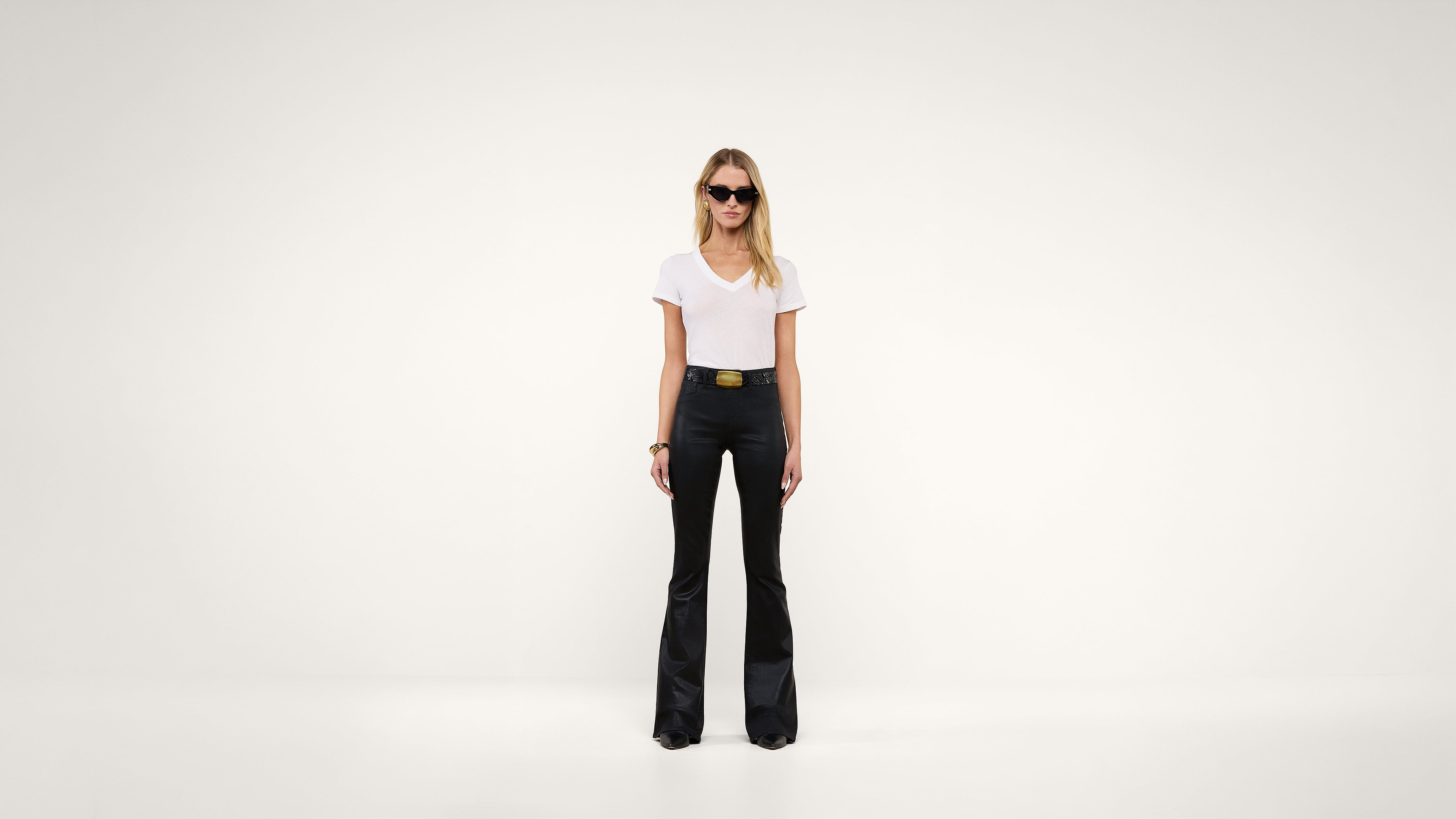 A woman wearing sunglasses, a white t-shirt, and black flared pants stands against a plain white background.