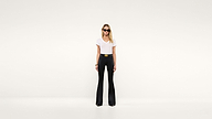 A woman wearing sunglasses, a white t-shirt, and high-waisted black flared pants stands confidently against a plain white background.