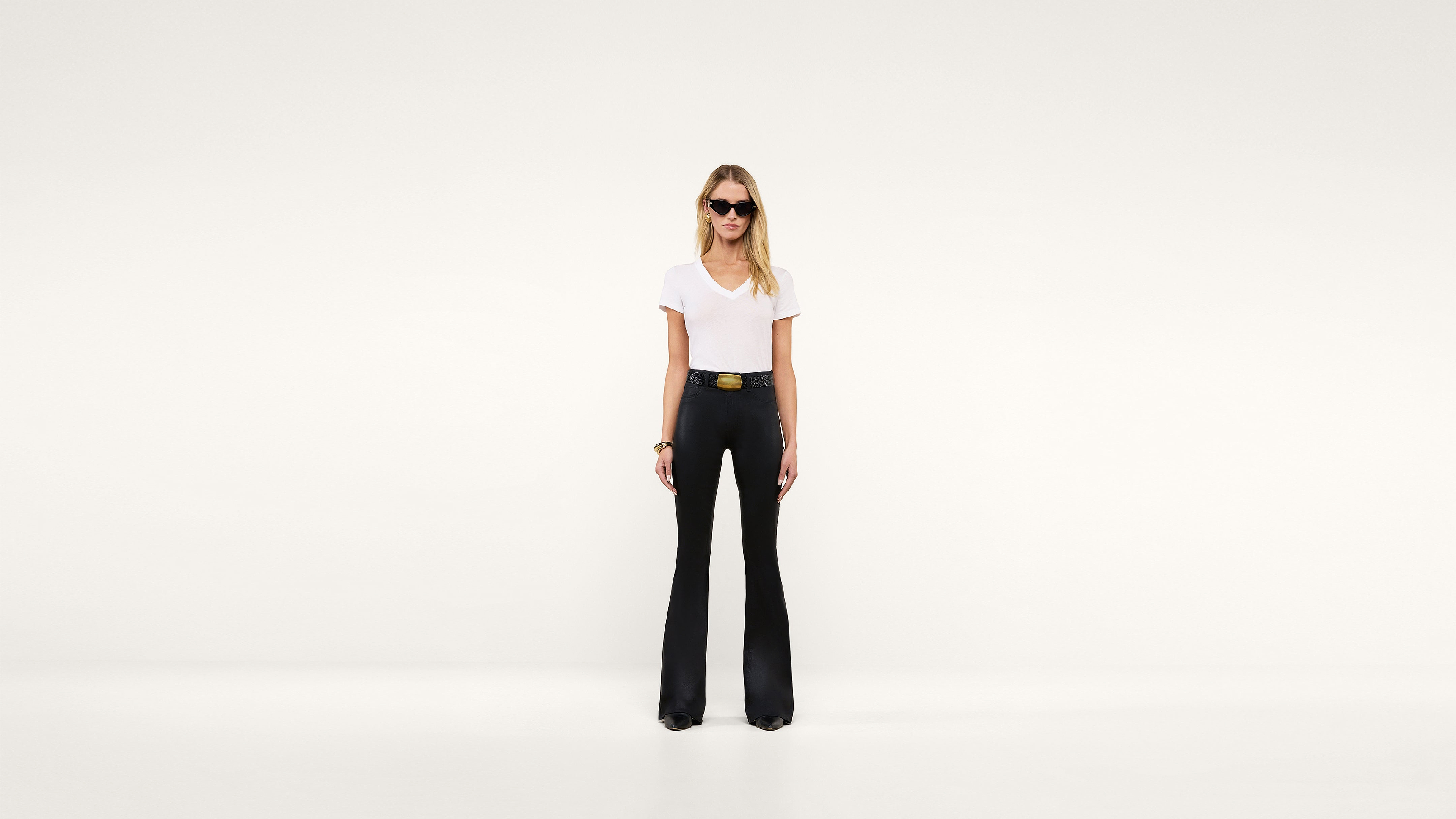 A woman wearing sunglasses, a white t-shirt, and high-waisted black flared pants stands confidently against a plain white background.