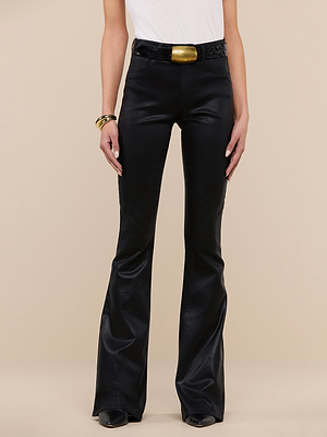 A person wearing Marty Coated Flare Jeans with a shiny finish, a white top, black belt with large gold buckle, gold bangles, and black shoes stands against a beige background.