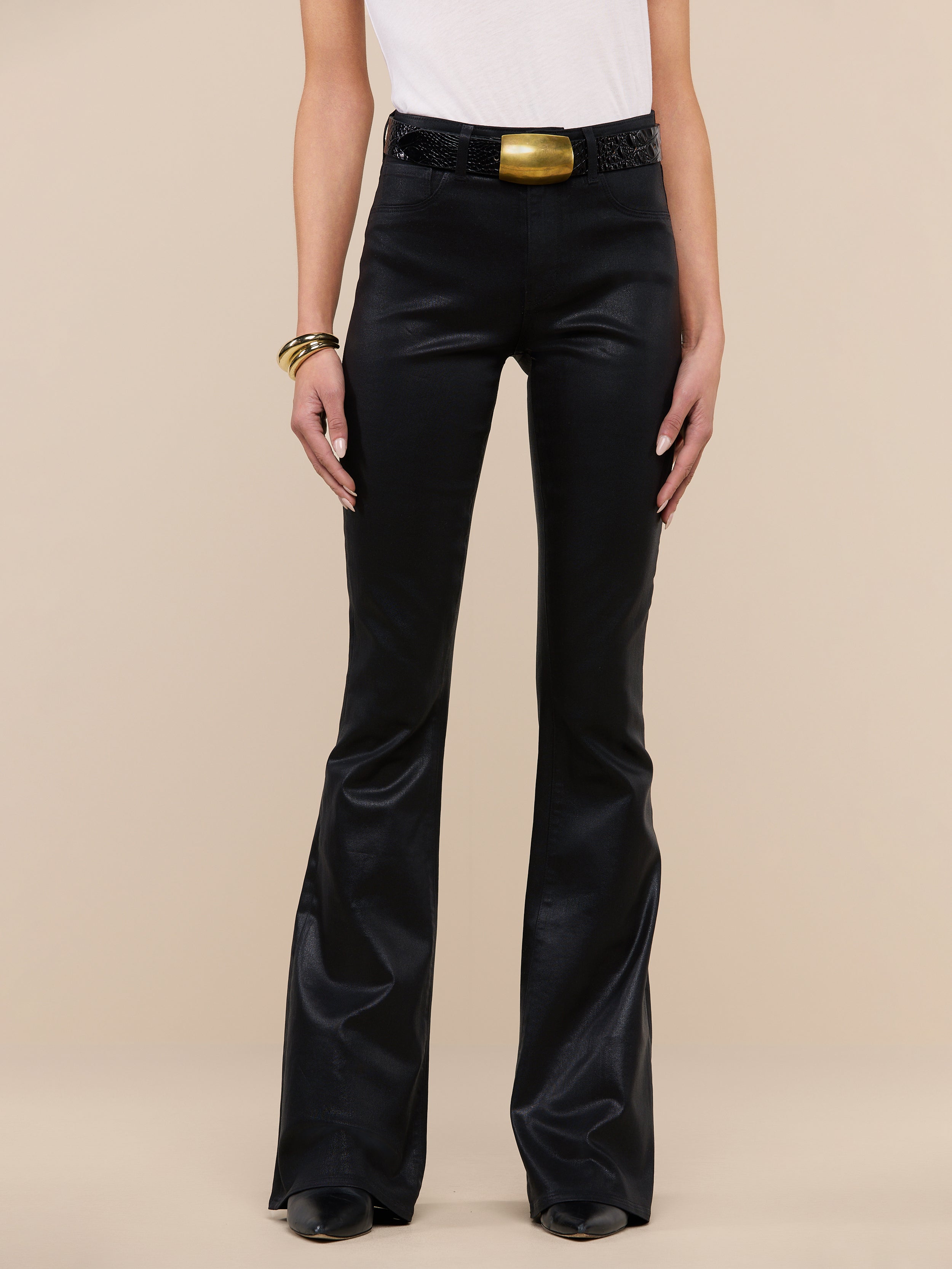 Wearing the Marty Coated Flare Jean in black, a person pairs the flared pants with a white sleeveless top, black shoes, a chunky belt featuring a gold buckle, and gold bracelets while standing against a plain white background.