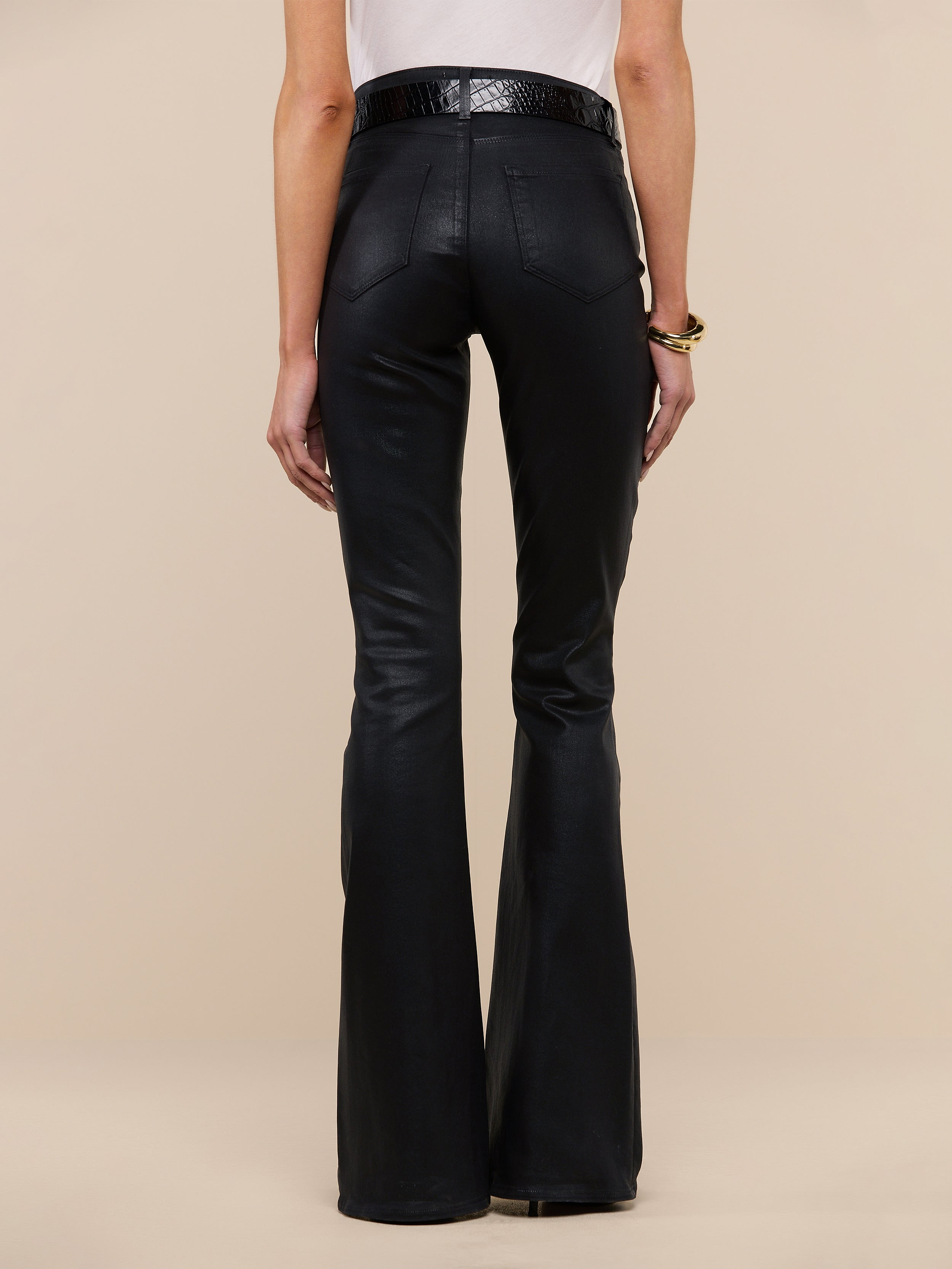 A person faces away from the camera, wearing a sleeveless white top and the Marty Coated Flare Jean in black, styled with a black belt. A bracelet adorns their left arm. The background is plain white.