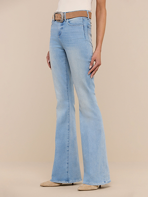 Wearing the Marty Flare Jean, styled with a beige belt and pointed-toe shoes, a person stands against a plain beige background. Only the lower torso and legs are visible.