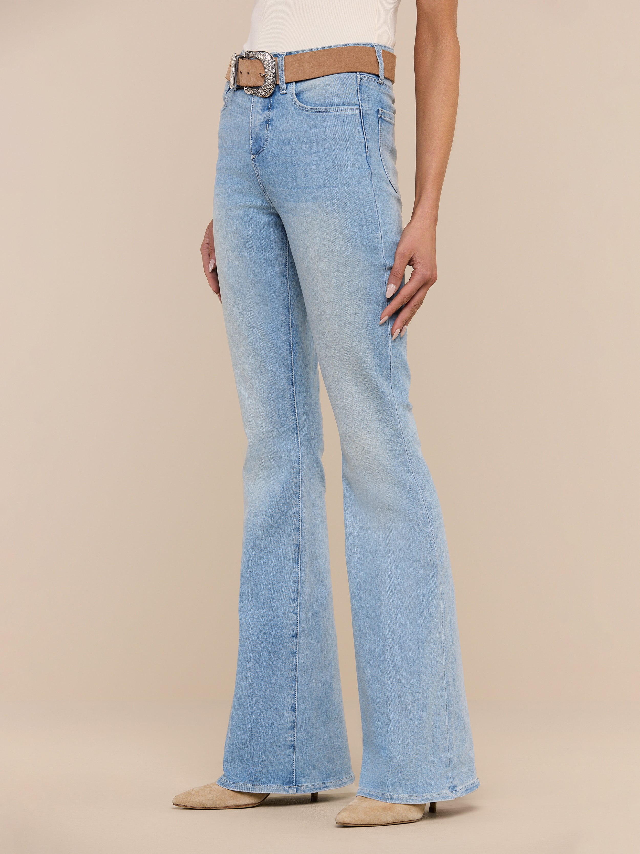 A person wearing the Marty Flare Jean in light blue, styled with a brown belt and beige pointed-toe shoes, stands against a plain white background. The upper body is mostly out of frame.