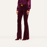A woman stands against a plain, light background, wearing the Marty Velvet Pant—high-rise, flare-leg pants in stretch burgundy velvet—paired with a matching long-sleeve blouse, brown belt, and pointed shoes. Her head is not visible in the image.