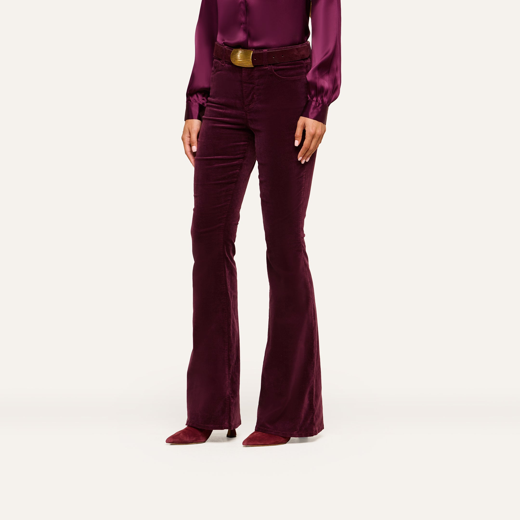 A woman stands against a plain, light background, wearing the Marty Velvet Pant—high-rise, flare-leg pants in stretch burgundy velvet—paired with a matching long-sleeve blouse, brown belt, and pointed shoes. Her head is not visible in the image.