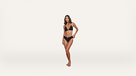 A woman stands barefoot against a plain white background, wearing a black bikini and posing with one hand on her hip.