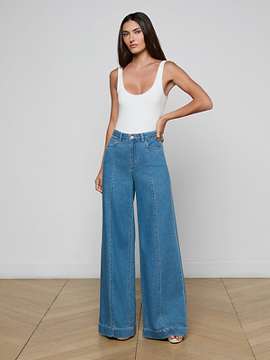 A woman with long straight hair and a neutral expression stands against a plain background wearing the Sandy Wide-Leg Jean in featherweight denim and a white sleeveless bodysuit, with one hand resting on her hip.