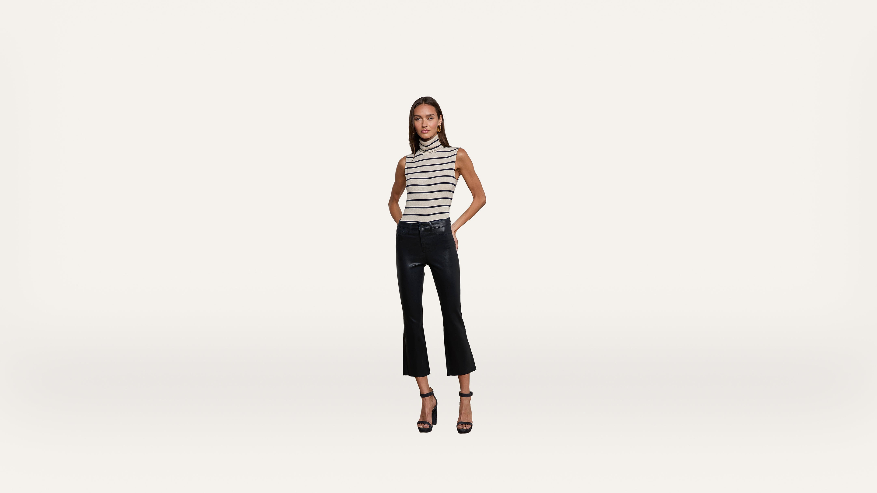 Woman in a sleeveless striped top, black cropped flared pants, and black heels stands against a plain white background, facing forward with hands in pockets.