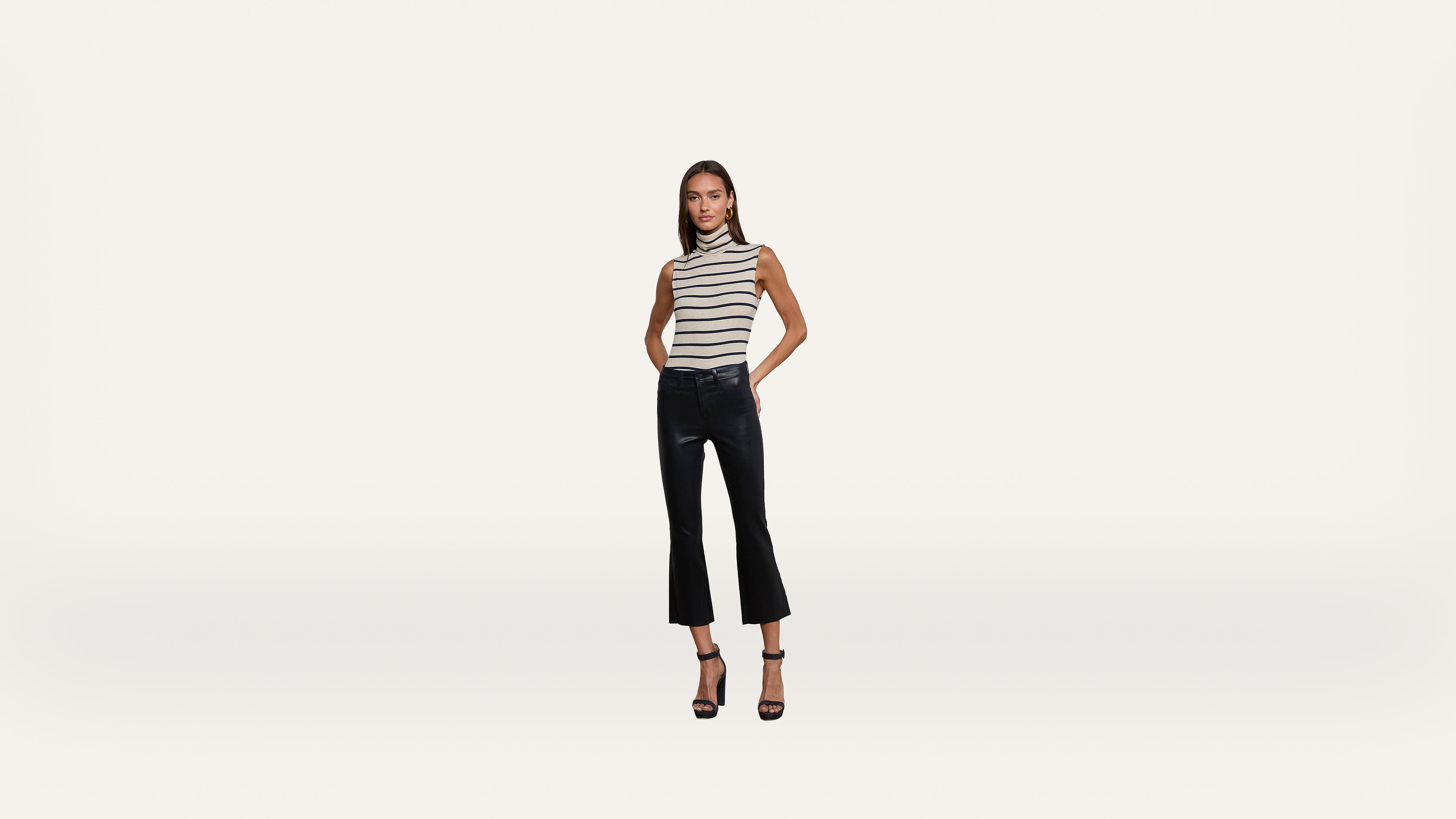 Woman in a sleeveless striped top, black cropped flared pants, and black heels stands against a plain white background, facing forward with hands in pockets.