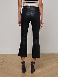 A person stands indoors on a wooden floor, facing away, wearing L'AGENCE Kendra Coated Cropped Flare Jeans, a sleeveless striped top, and black open-heeled shoes against an off-white wall.