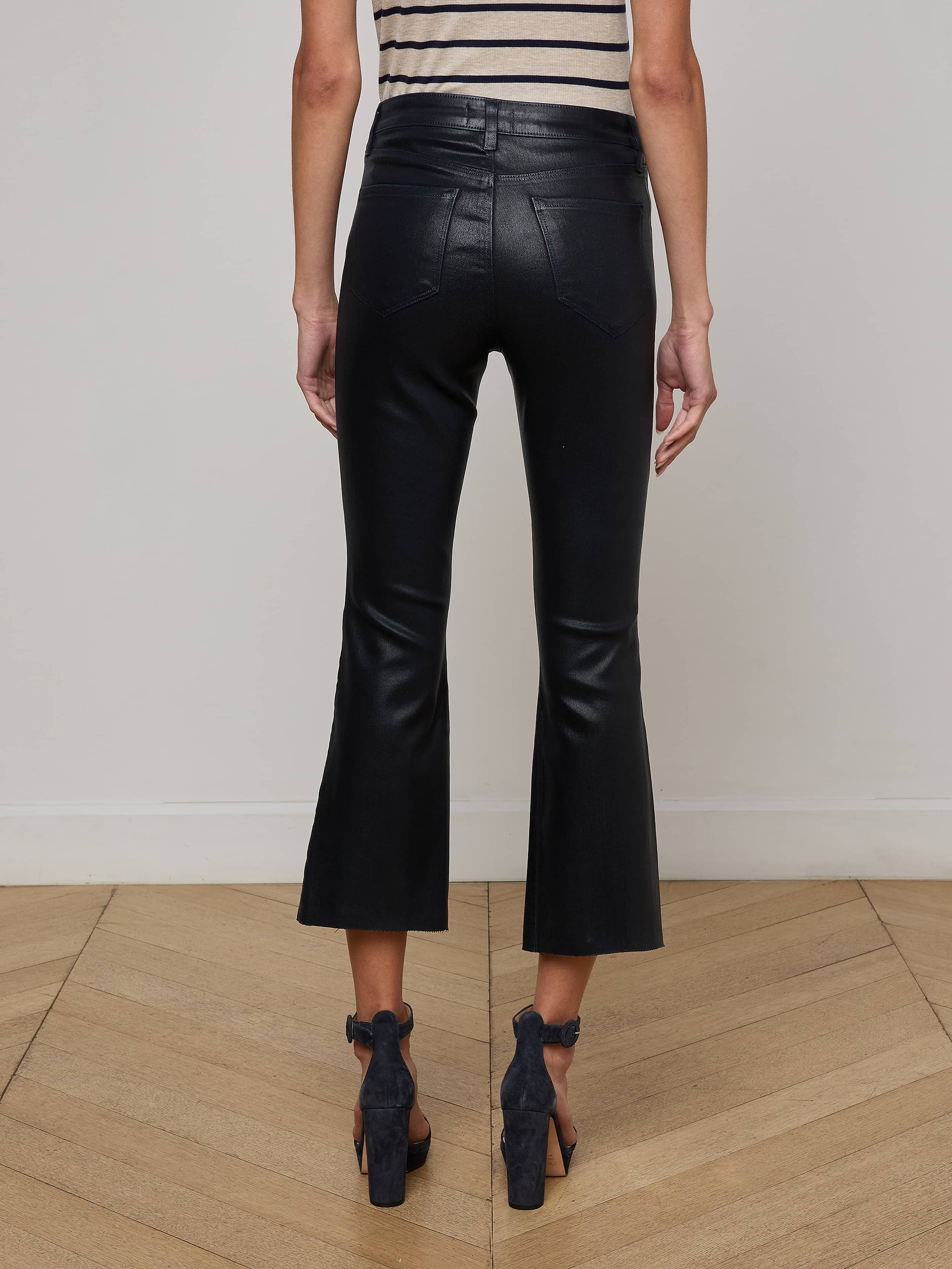 A person stands indoors on a wooden floor, facing away, wearing L'AGENCE Kendra Coated Cropped Flare Jeans, a sleeveless striped top, and black open-heeled shoes against an off-white wall.