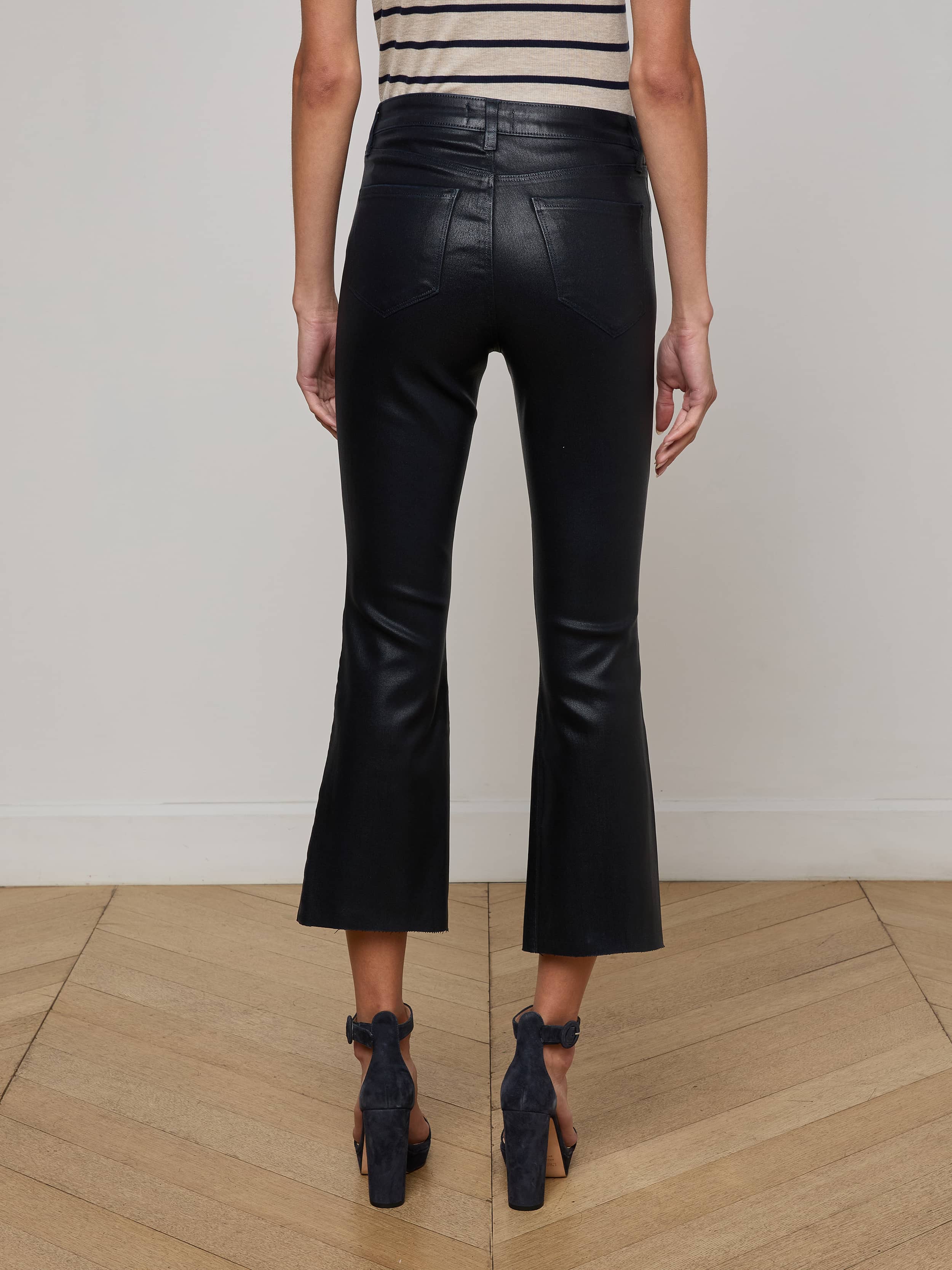 A person stands indoors on a wooden floor, facing away, wearing L'AGENCE Kendra Coated Cropped Flare Jeans, a sleeveless striped top, and black open-heeled shoes against an off-white wall.