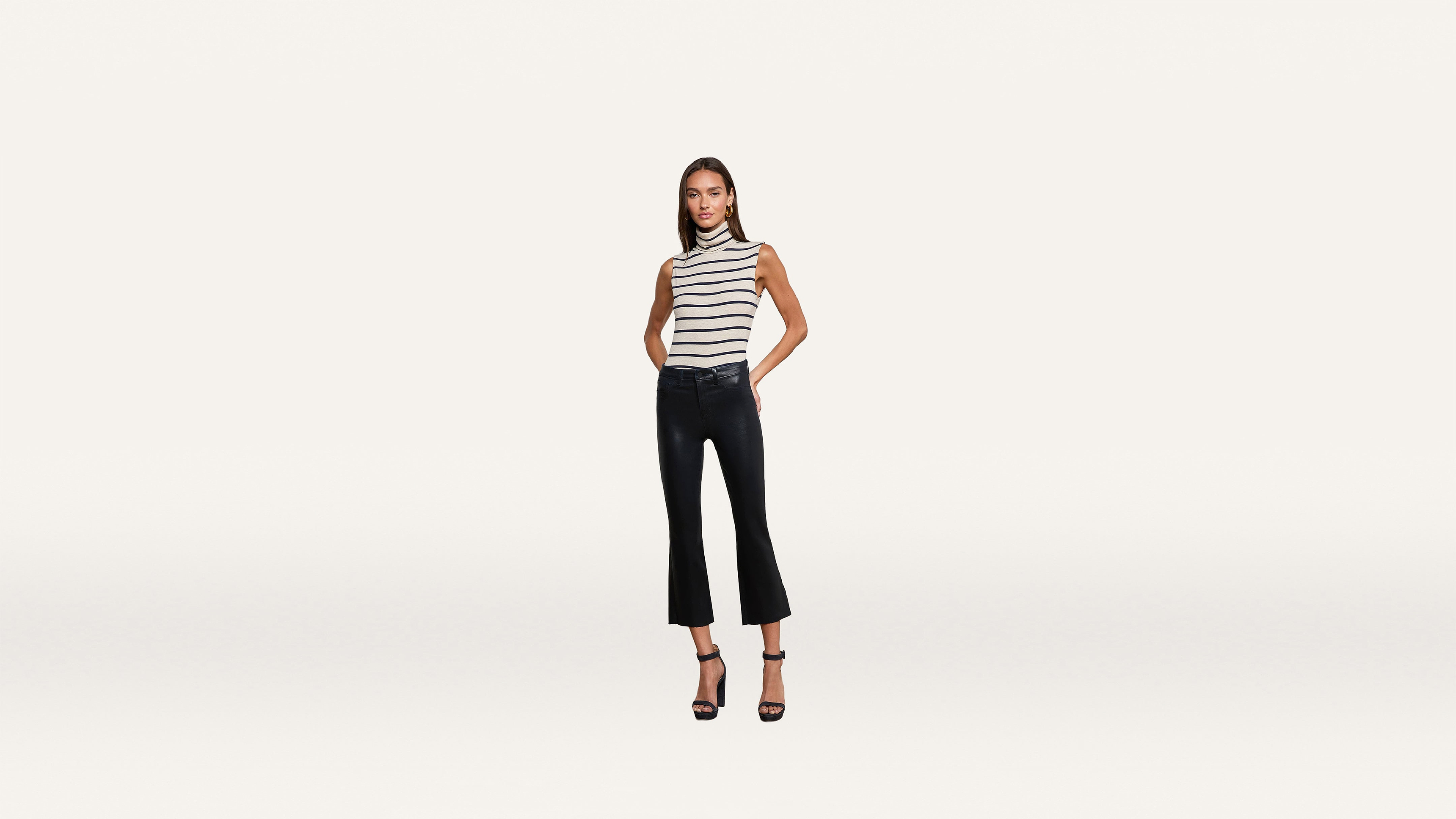 A woman stands against a plain white background wearing a sleeveless black-and-white striped top, black cropped pants, and stylish black high-heeled sandals.