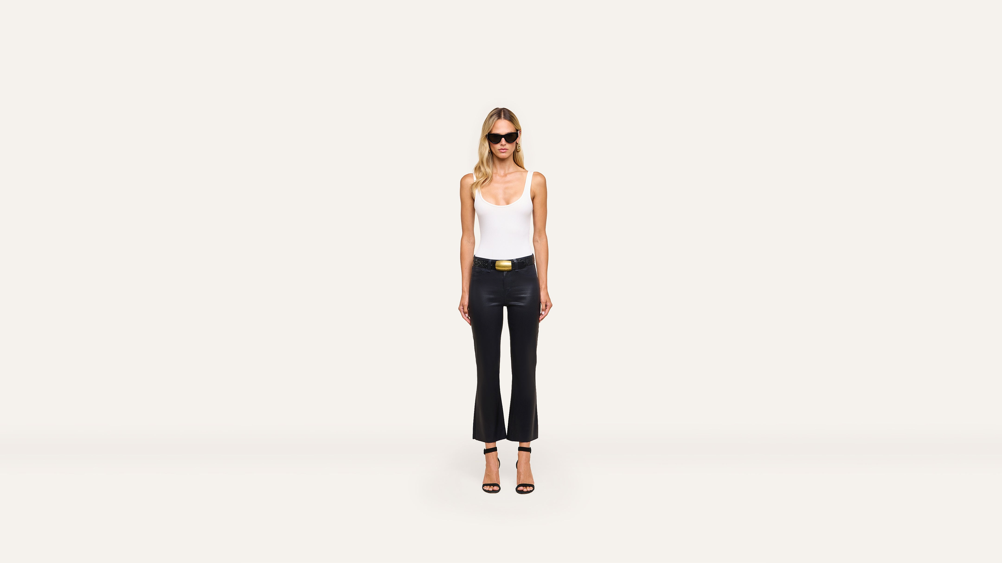 A woman wearing sunglasses, a white tank top, black pants, and heels poses confidently against a plain white background.