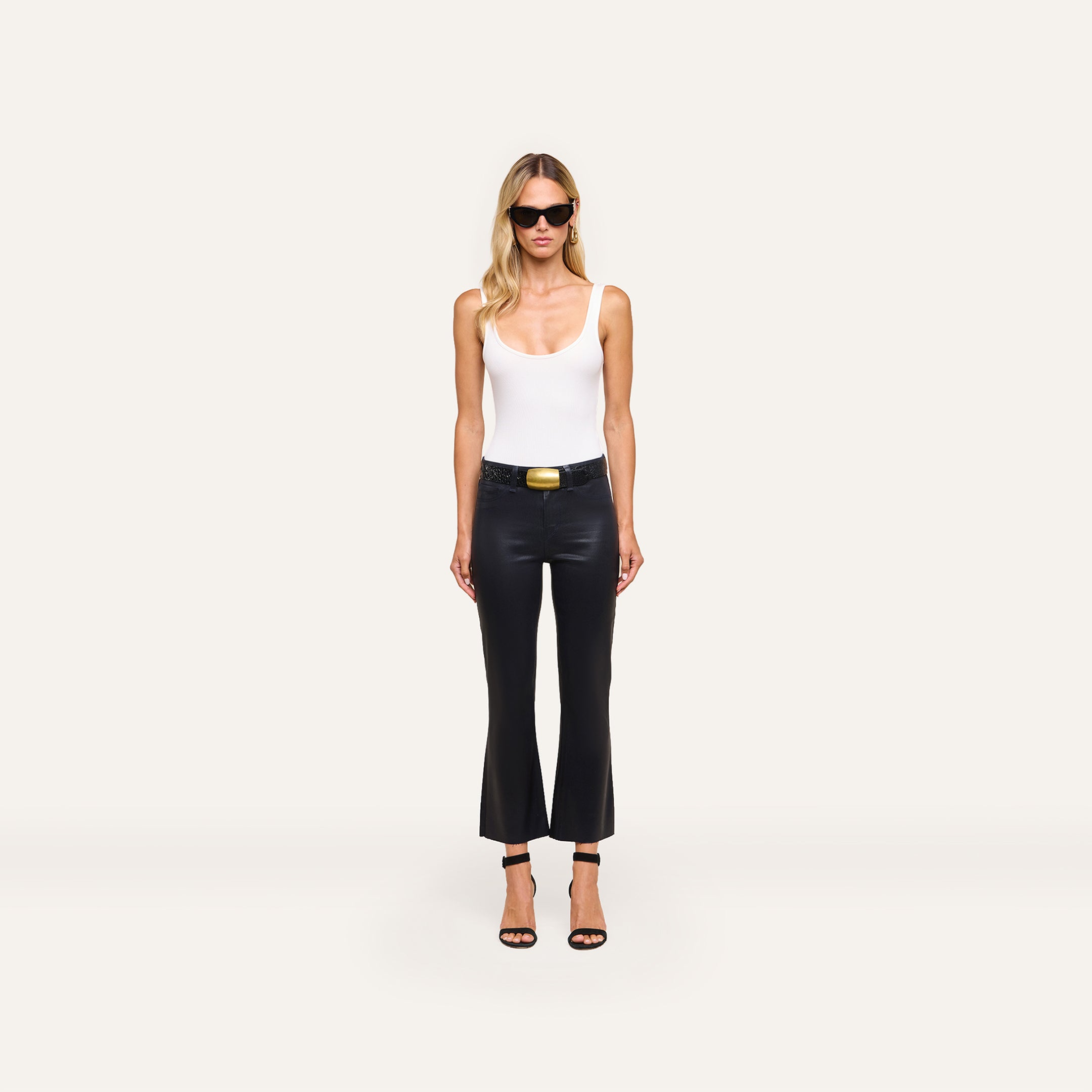 A woman in sunglasses, a white tank top, the Kendra Coated Cropped Flare Jean with a belt, and black heels stands against a plain white background.