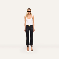 A woman in sunglasses, a white tank top, the Kendra Coated Cropped Flare Jean with a belt, and black heels stands against a plain white background.