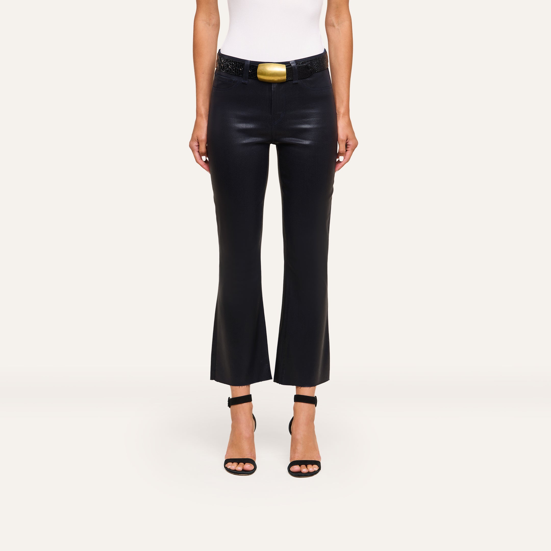 A person wears the Kendra Coated Cropped Flare Jean, a white sleeveless top, a black belt with a large gold buckle, and black heeled sandals, standing against a plain light background.