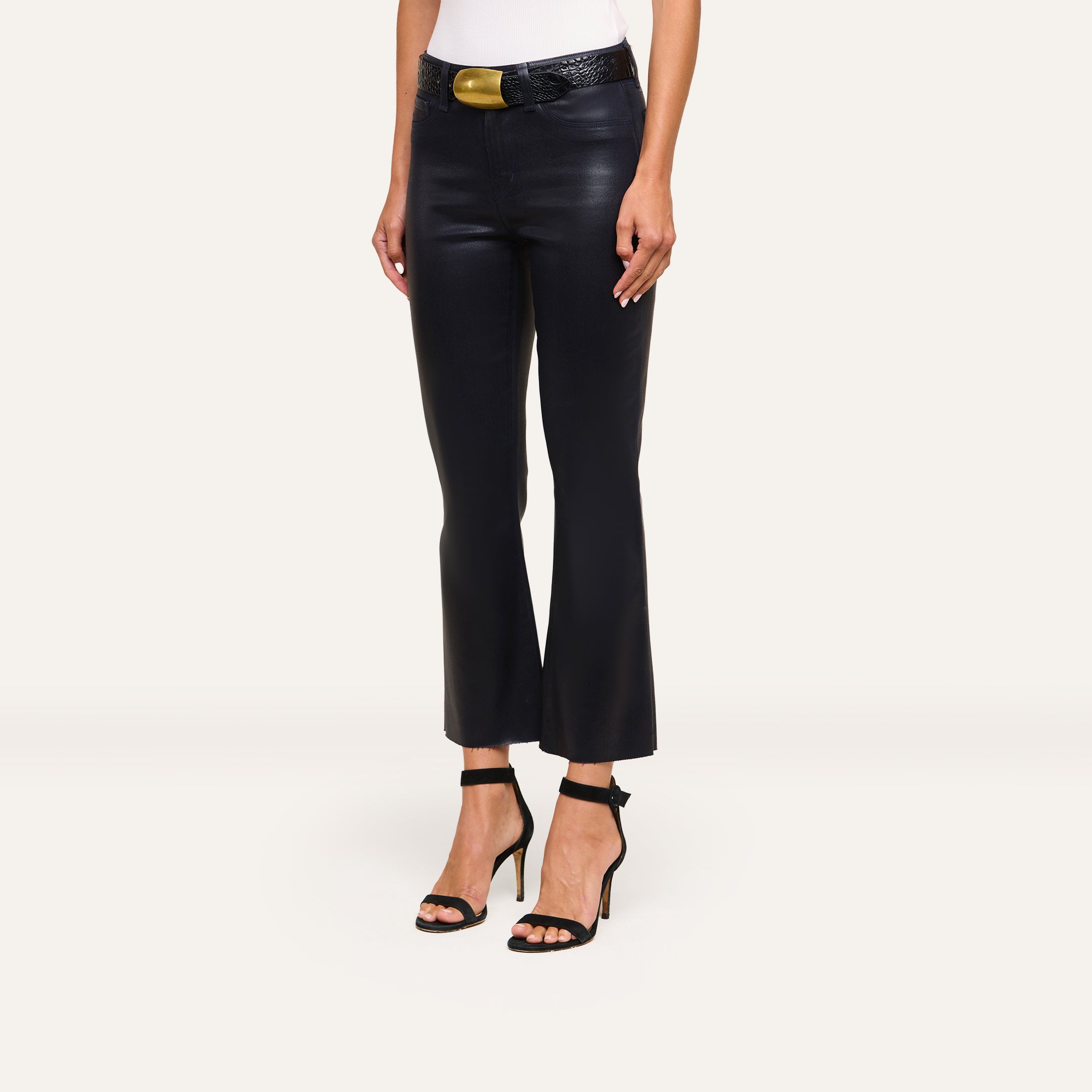 A person wears Kendra Coated Cropped Flare Jean, a white top, a black belt with a gold buckle, and black high-heeled sandals, standing against a plain light background.